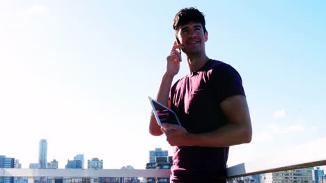 Man-talking-on-mobile-phone-while-holding-digital-tablet-in-balcony-4k