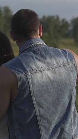close backside view muscular man in denim vest hugs brunette lady and shows with hand against green trees