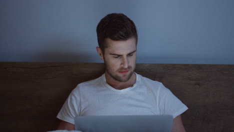 caucasian man in white t-shirt working on the laptop late at night while sitting in the bed before sleeping