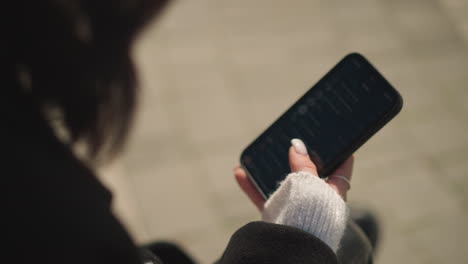 close-up of lady with short hair wearing black coat and white sweater scrolling on smartphone with focus on hand holding phone showing visible manicure