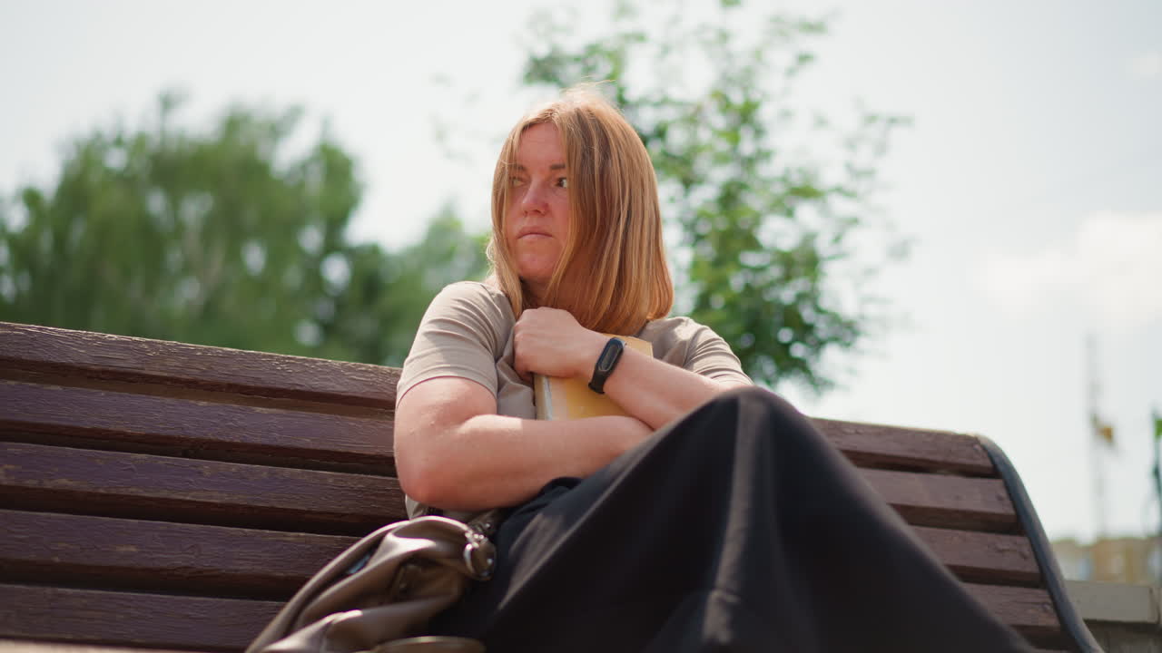 Thoughtful woman sitting on wooden bench outdoors holding closed book tightly to chest, looking upward with reflective calm expression under bright daylight, trees in background