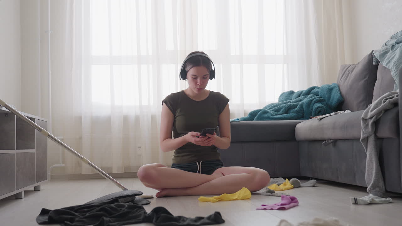 Lady seated cross legged wearing headphones listening to music while operating phone in messy living room with mop and scattered clothes on floor near sofa under sunlight filtering through curtain