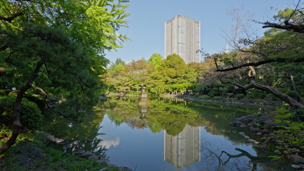 A tranquil pond in Hibiya Park, reflecting lush green trees and a prominent tall city building under a clear blue sky.