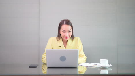 Elegante-Mujer-De-Negocios-Asiática-Sentada-En-El-Escritorio-Frente-A-Su-Computadora-Portátil-Y-Escribiendo-En-El-Teclado-Con-Una-Expresión-De-Cara-Feliz,-Vista-Frontal-En-Cámara-Lenta