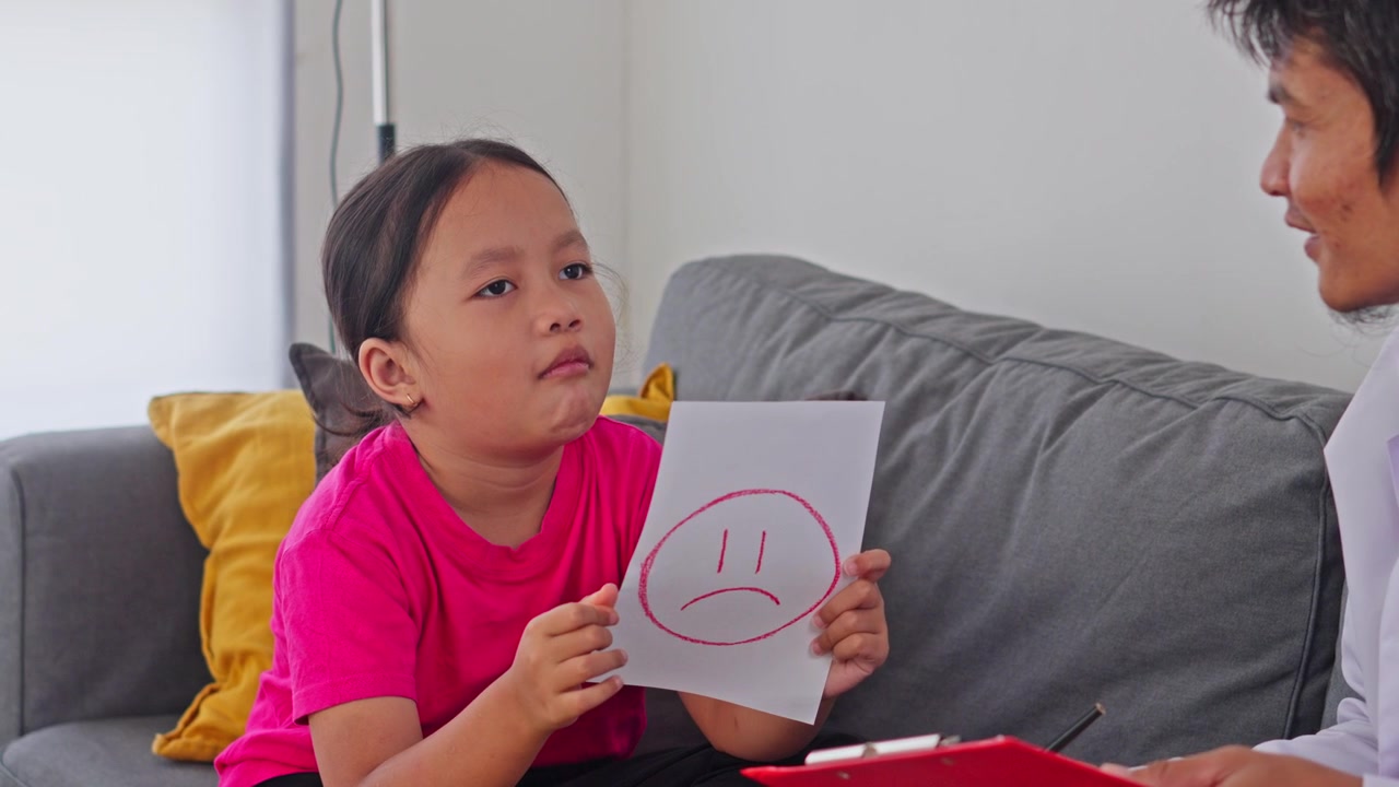 Bambina asiatica che tiene in mano un disegno con una faccia triste e parla con la sua psicologa durante la terapia