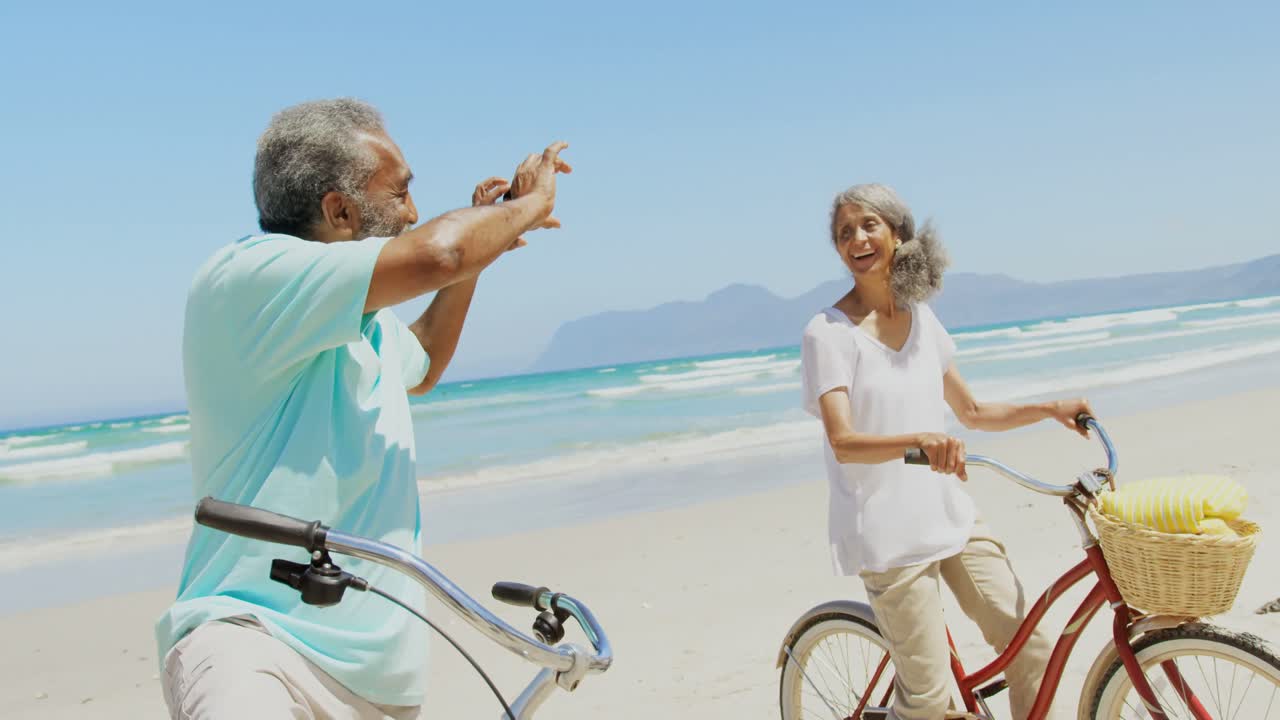 Side view of active senior African American man taking photo of woman with mobile phone at beach 4k