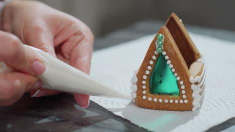 close-up of hands carefully piping white icing onto gingerbread house with emerald green sugar window, placed on textured tissue paper, showcasing intricate details and festive decoration