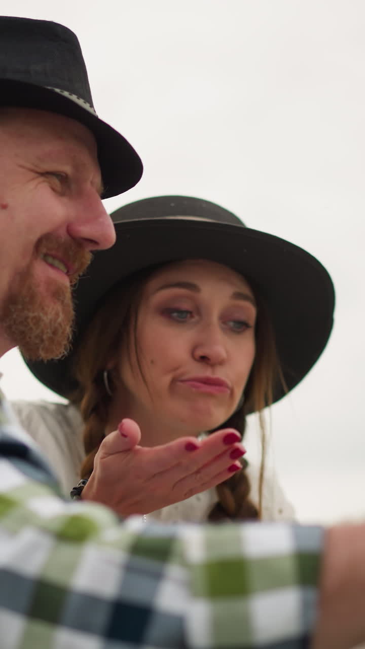 se ve a un artesano con una camisa a cuadros pintando felizmente en una tabla en un entorno al aire libre sereno y verde. a su lado, una mujer con un vestido blanco y un sombrero de pie, su expresión ligeramente no convencida