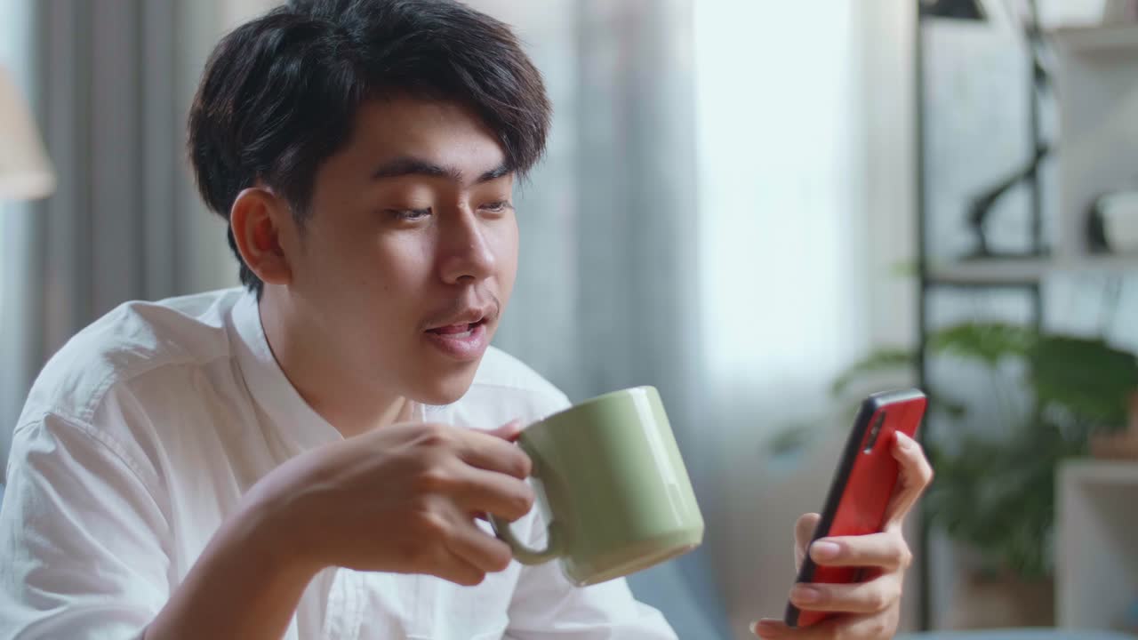 Close Up Side View Of Happy Asian Man Drinking Coffee While Using Smartphone In The Living Room
