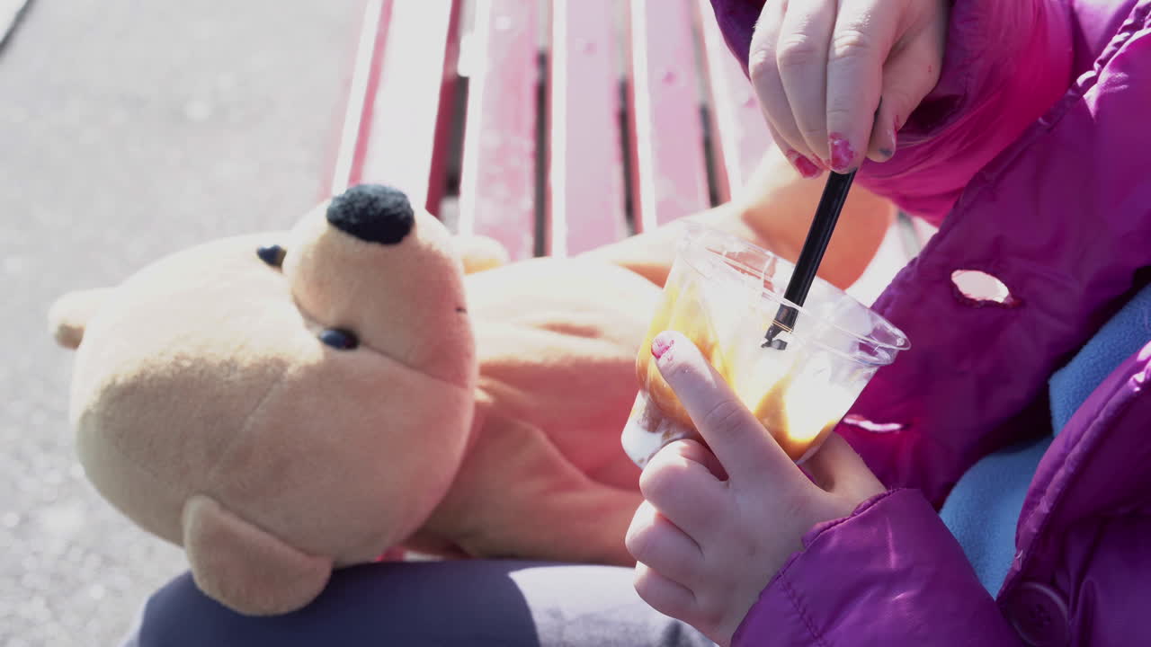 파란색 점퍼 매니큐어와 테디베어를 들고 야외에 앉아 있는 선디 아이스크림을 들고 있는 어린 소녀의 클로즈업