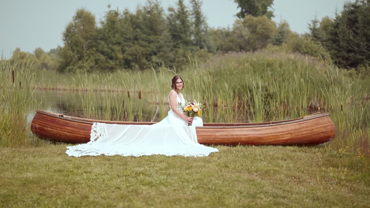 Slow-motion shot of bride in canoe; she looks up and smiles, bouquet in hand
