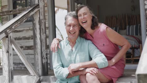 Portrait-of-happy-senior-caucasian-couple-embracing-sitting-on-steps-of-beach-house,-in-slow-motion