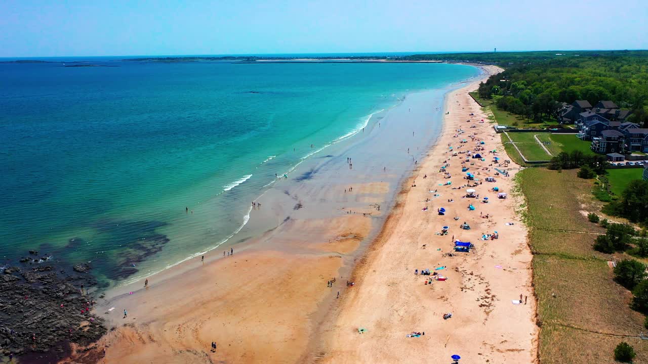 Aerial drone view of a lively Saco, Maine beach. People swim, sunbathe, and relax while waves crash on sandy shore. Vacation homes line the coast, adding charm to this peak-season East Coast vibe