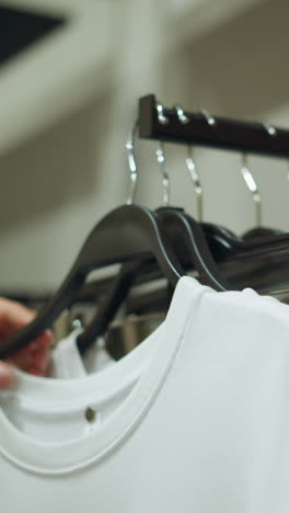 una mano en tela rosa devuelve cuidadosamente una tapa blanca a un estante de ropa con pantalones de ceniza, el fondo muestra una vista borrosa de ropa bien arreglada en los estantes con un entorno de tienda bien iluminado