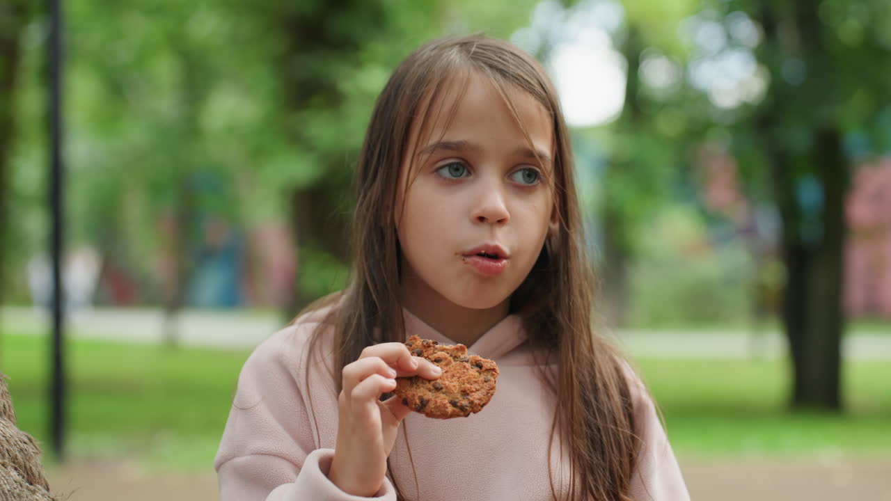 Child enjoying snack. Child with pink hoodie munching cookie in peaceful park setting. Young girl wearing pink examines her cookie carefully while sitting on swing in tranquil green surroundings