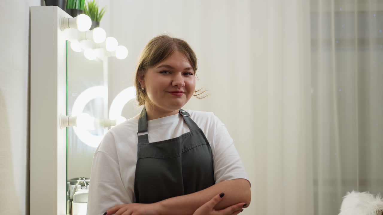Young woman with short blond hair smiling softly while standing with folded arms in stylish salon interior, featuring glowing ring light, vanity mirror with bulbs, and white curtains