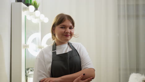young woman with short blond hair smiling softly while standing with folded arms in stylish salon interior, featuring glowing ring light, vanity mirror with bulbs, and white curtains