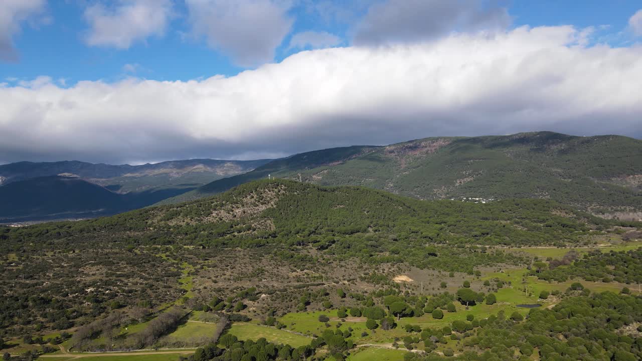 un vuelo retrocediendo en un valle con un sistema montañoso en primer plano, sus picos cubiertos por impresionantes nubes, descubriendo bosques de pinos y prados verdes en una soleada mañana de invierno en ávila, españa