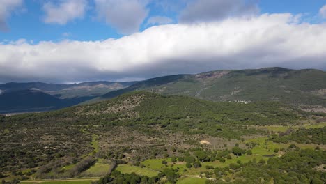 receding-flight-in-a-valley-with-a-mountain-system-in-the-foreground,-its-peaks-covered-by-impressive-clouds,-discovering-pine-forests-and-green-meadows-on-a-sunny-winter-morning-in-Avila-Spain