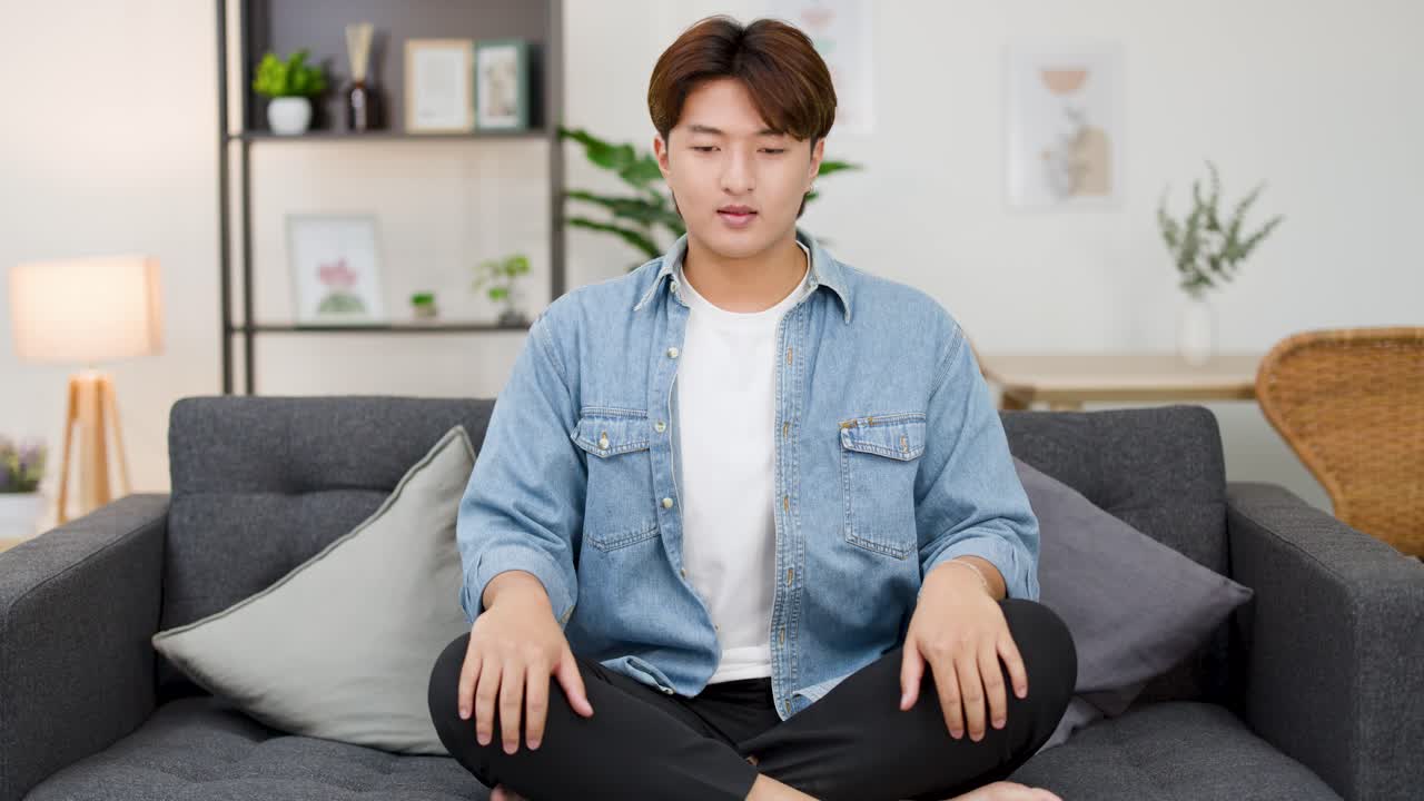 明るい部屋で、若いアジア人男性がソファに胡坐をかいて座り、目を閉じてマインドフルネスを実践しています。