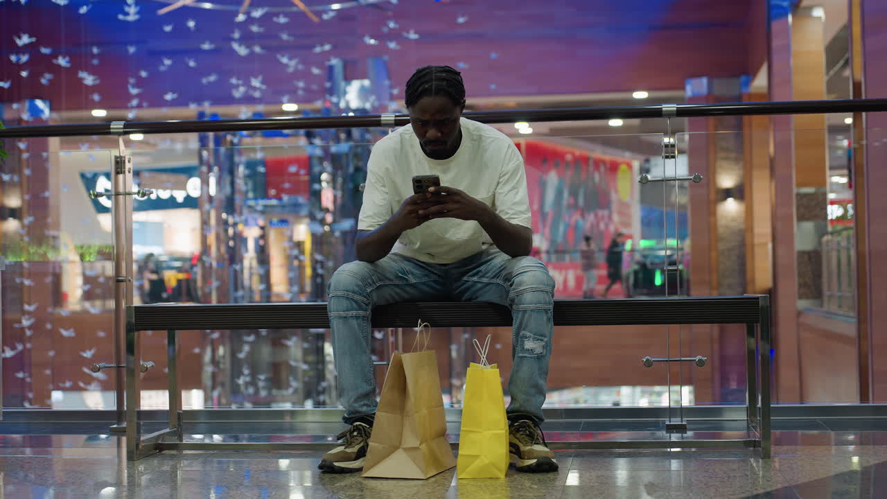 Young black man in white shirt and ripped jeans sits on bench with brown and yellow shopping bags, fully focused on phone, while colorful modern mall and blurred shoppers decorate background
