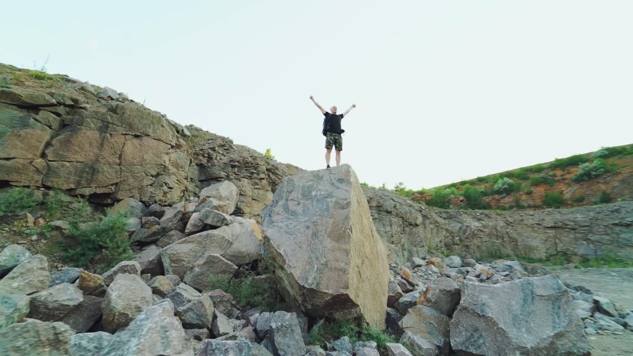 happy traveler with a backpack on his shoulders is standing at the top of a cliff with his arms raised and looking at the beautiful nature from high. Camera motion back