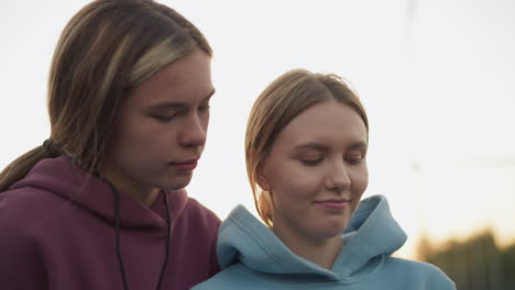 close-up of two women, one in maroon hoodie showing something to other in blue hoodie, with a warm interaction in outdoor volleyball court setting