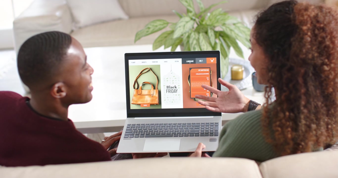 African American couple on sofa browsing Black Friday laptop, comparing handbag vs phone to decide