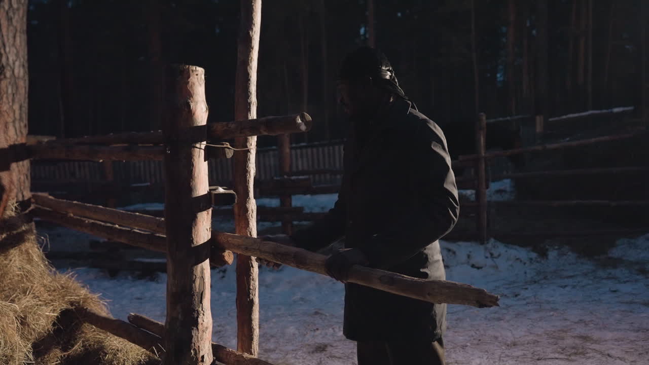 울타리 옆 목재 실루엣, 겨울 황혼에 목재를 운반하는 모습, 추운 저녁 햇살 아래 눈 덮인 마당을 가로질러 울타리 쪽으로 나무 레일을 운반하는 사람