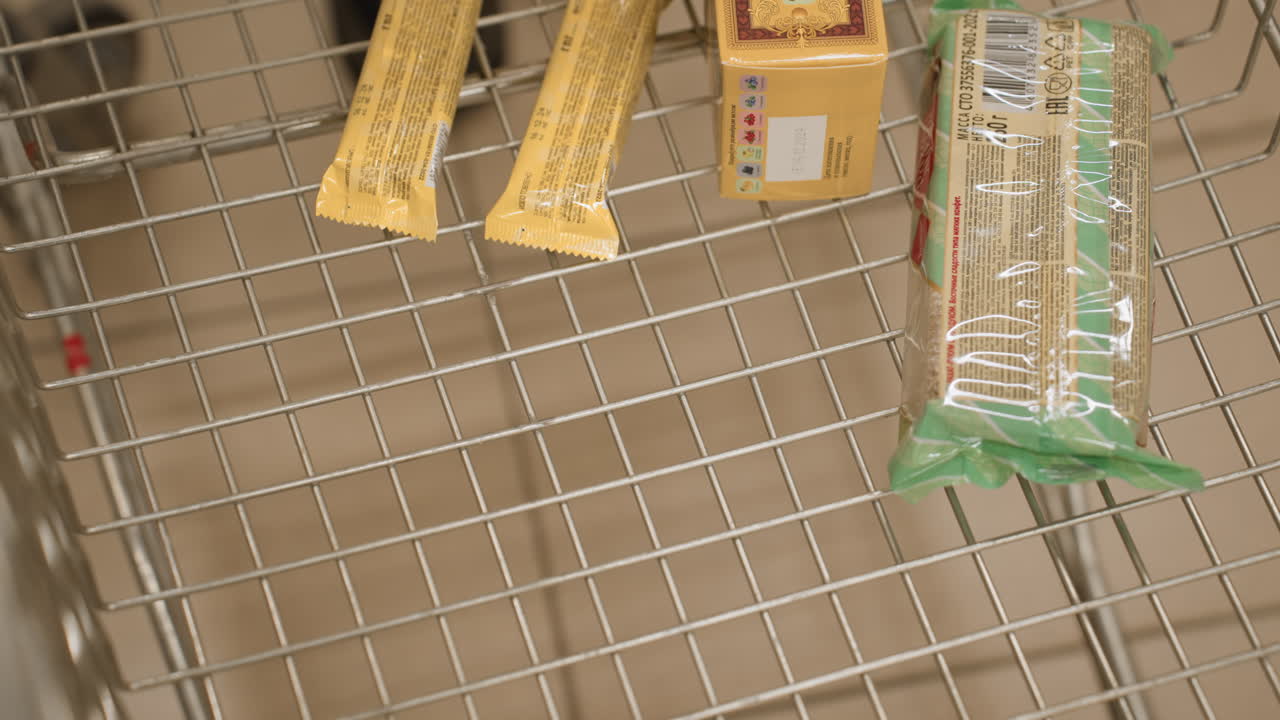 Close up of shopper dropping pink cupcake box with swirl design into metal trolley in supermarket aisle, highlighting treat selection and motion of hand placing sweet bakery item into cart