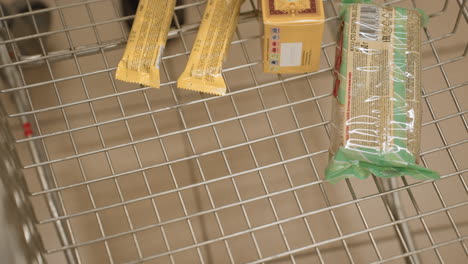 close up of shopper dropping pink cupcake box with swirl design into metal trolley in supermarket aisle, highlighting treat selection and motion of hand placing sweet bakery item into cart