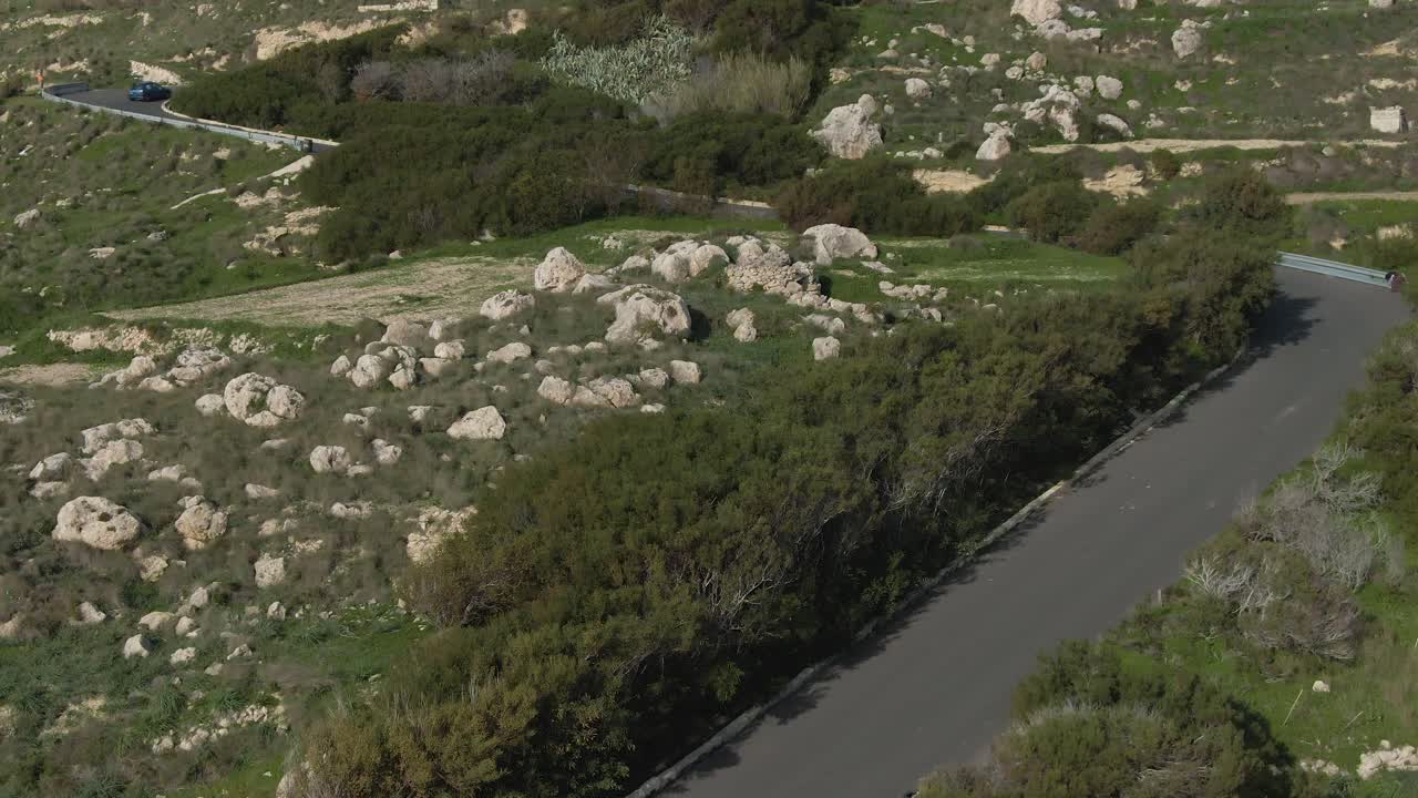 Blue Car Driving Fast On The Road Between The Lush Fields With Green Trees And Rocks On A Hot Sunny Weather - Aerial Shot