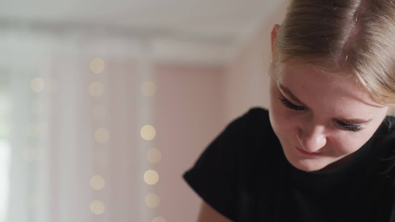 Close-up of light skin woman wearing black shirt and smiling gently while looking down, soft indoor lighting creates warm mood with out-of-focus pastel wall background adding calm ambiance