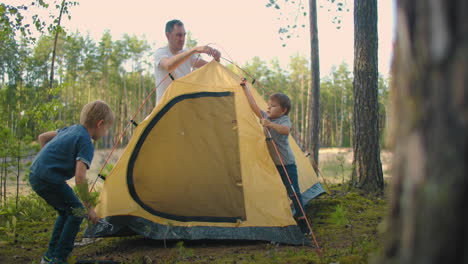 the children together with their father set up a tent for the night and camping in the forest during the journey. a man and two children 3-5 years old together in a hike gather a tent in slow motion