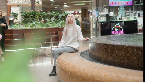 long shot of mindful explorer woman seated close to mosaic water fountain in busy mall hall with neon signage, ambient light and moving people in background, gazing thoughtfully around