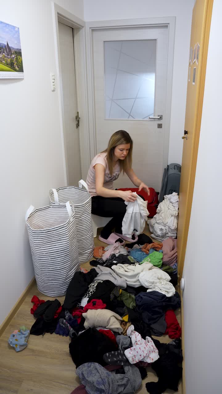 Incombenza post-viaggio: donna che sistema una grande pila di vestiti sporchi sul pavimento del corridoio con cesti della biancheria nelle vicinanze. Disfare i bagagli, pulizia e ripristino della casa dopo un viaggio. Verticale.