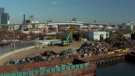 Slide-and-pan-footage-of-loader-moving-on-scrap-yard-on-Newtown-Creek-waterfront.-Piles-of-used-metal-material-for-recycling.-Busy-elevated-road-in-background.-New-York-City,-USA