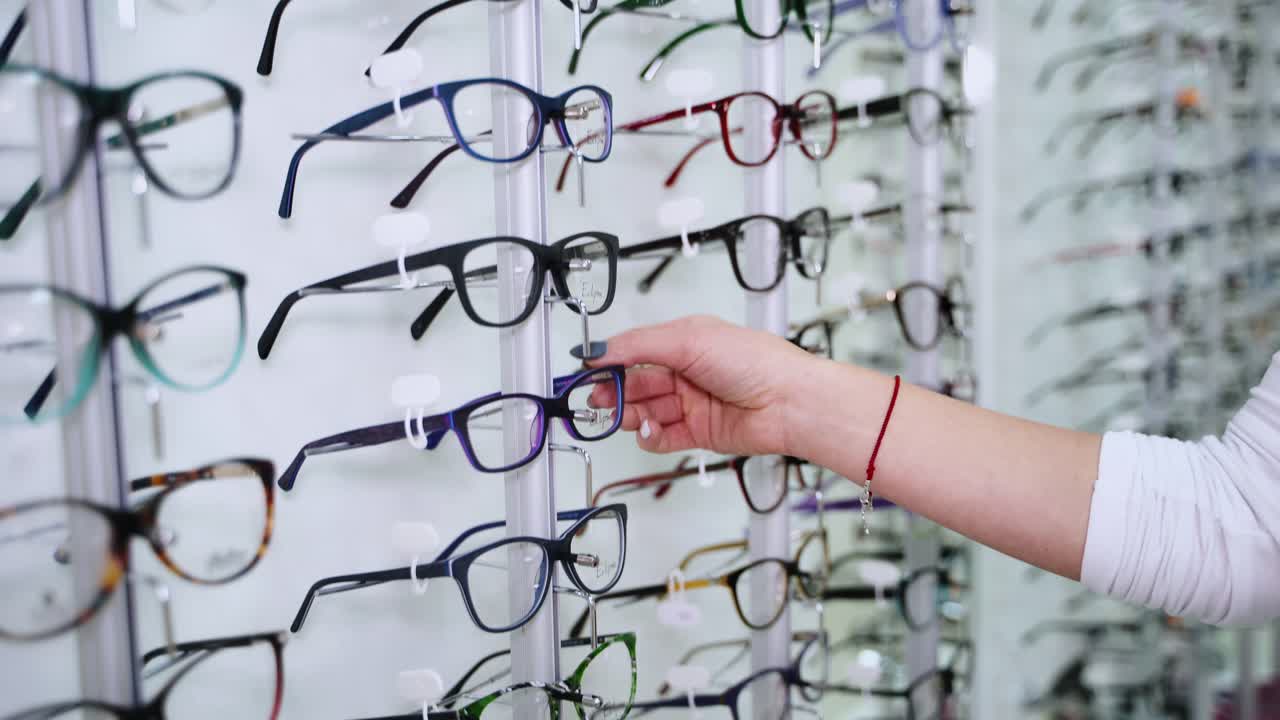 Woman's hand choosing eyeglasses on the stand indoors. Female is taking new glasses at the optic store.