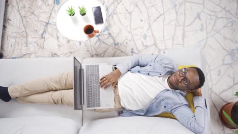 Thoughtful-happy-african-young-man-resting-on-sofa-at-home-and-using-laptop.