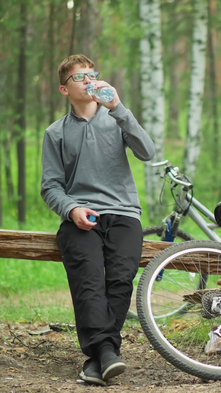 un niño joven con gafas y una camiseta de manga larga gris toma un sorbo de agua mientras está sentado en un tronco con la pierna cruzada, descansando al lado de una bicicleta estacionada en el bosque, rodeado de altos árboles verdes