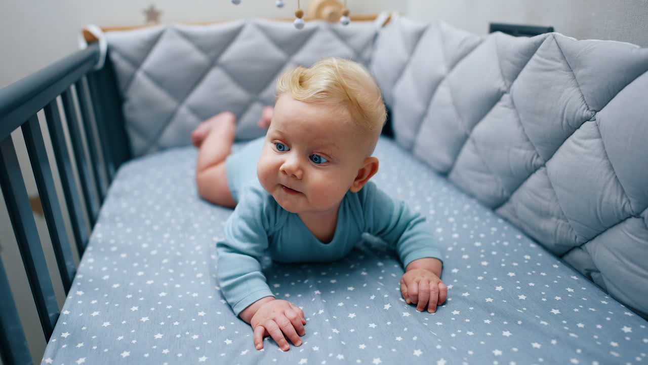 Cute blue-eyed baby boy in blue bodysuit lies on his belly. Adorable child in his cozy crib.