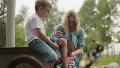 a mother squats as she carefully cleans her son's bleeding leg with a tissue while the boy holds his pant leg up