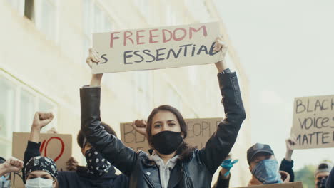 mujer caucásica con máscara facial sosteniendo un cartel de "la libertad es esencial" en una protesta con un grupo multiétnico de personas en la calle"