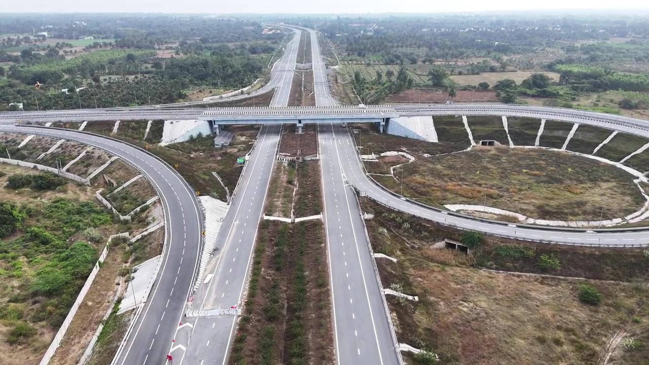 Drone shot capturing a modern expressway junction with curved connectors and structured overpasses, showcasing large-scale roadway engineering surrounded by agricultural terrain and scattered greenery