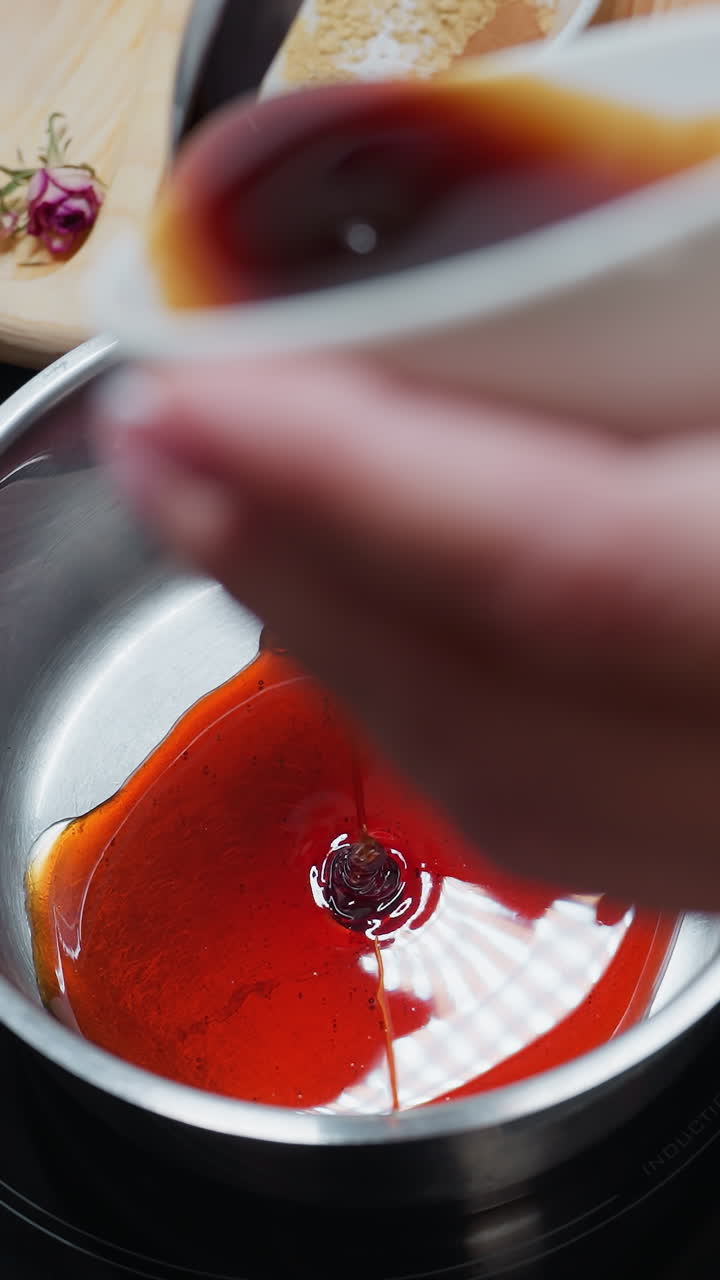 vista de mano de primer plano de una persona vertiendo cuidadosamente miel de un pequeño cuenco blanco en una olla en una estufa eléctrica, encimera de la cocina con ingredientes en el fondo, iluminación cálida que crea una atmósfera acogedora