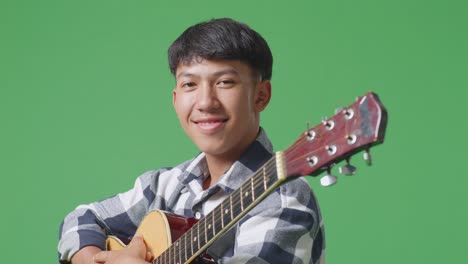primer plano de un adolescente asiático con una guitarra cruzando los brazos y sonriendo en el fondo de la pantalla verde
