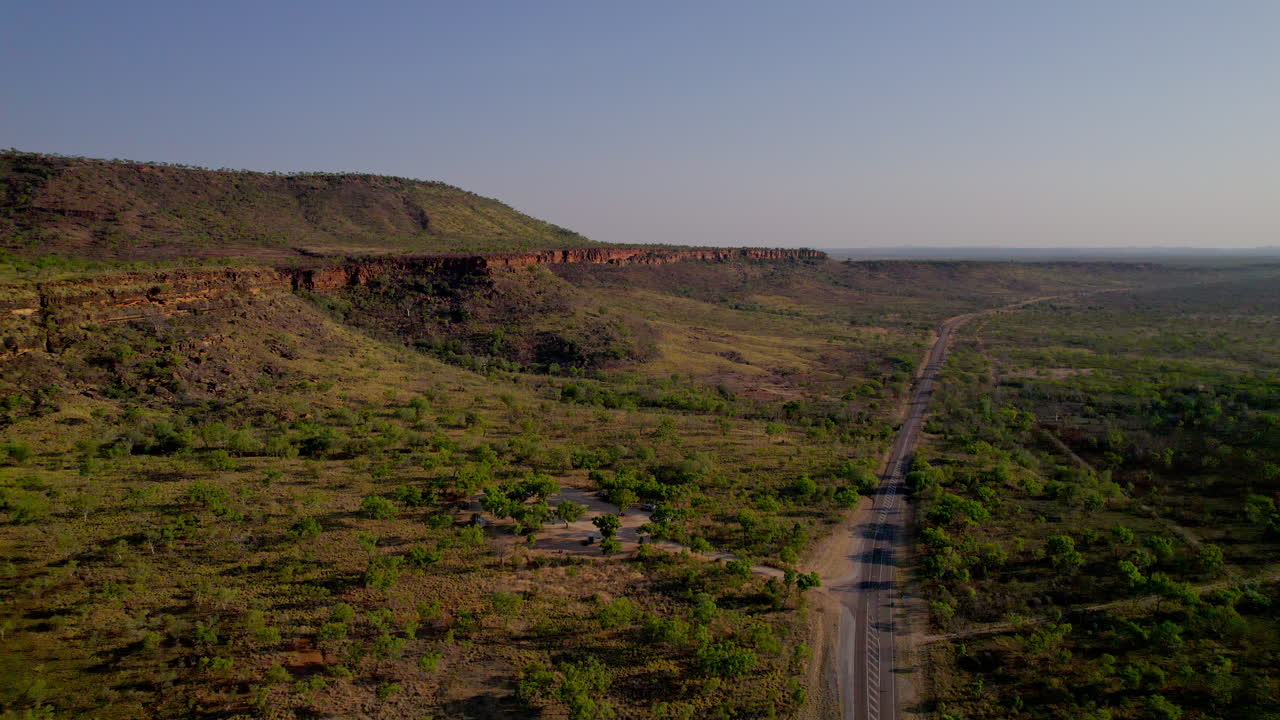 Un drone vola lungo la Victoria Highway verso l'Area di Sosta Saddle Creek nel Parco Nazionale Gregory, Territorio del Nord, rivelando colline rocciose, falesie rosse e boscaglia nella calda luce serale.