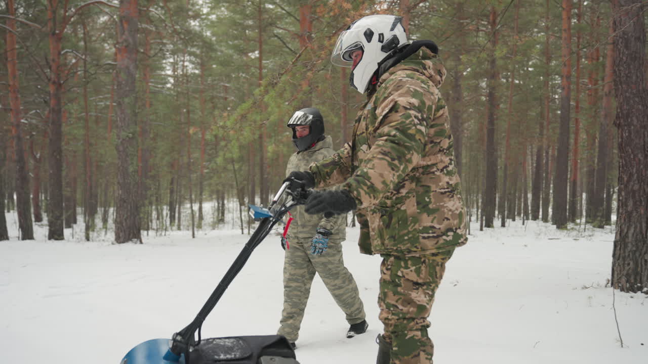 Snow pine forest atv prep helmeted rider adjusts controls on blue tracked atv while companion observes, camouflage gear, light snowfall, closeup on track and skis, cautious testing and equipment setup