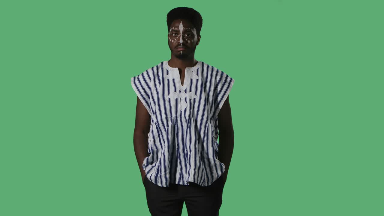 Portrait young African American man seriously looking at the camera. Black male with an ethnic white pattern on face and in striped national shirt posing on green screen in studio