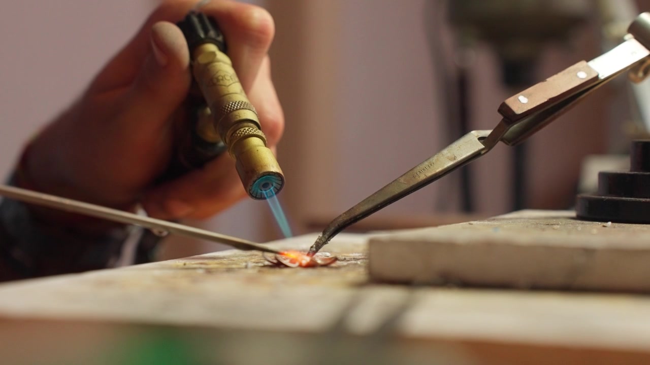 A craftsman is focused on using a torch to solder metal pieces together, highlighting precision and skill in a well-equipped workshop setting during the day.